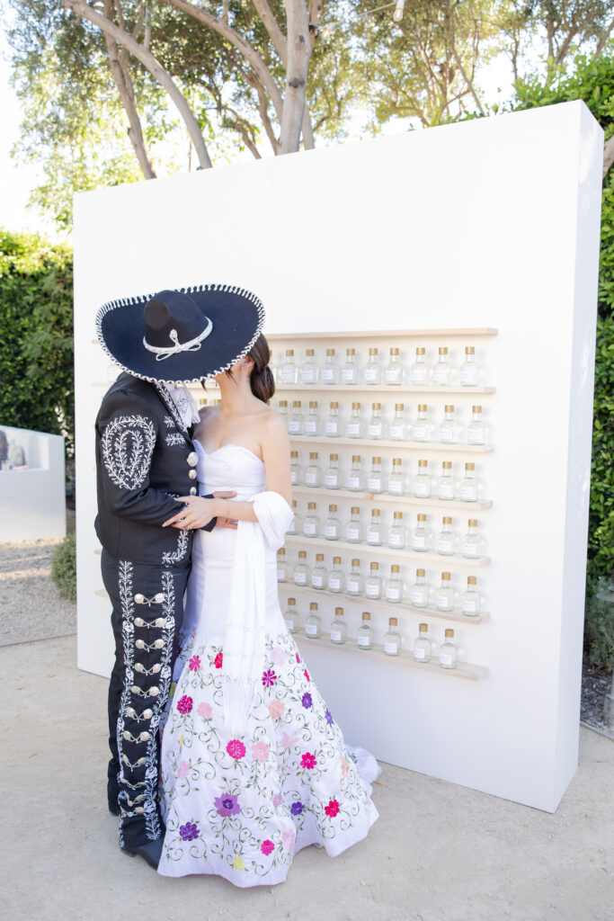 Bride and groom dressed in traditional Mexican wedding attire kissing in front of a white seating chart wall.