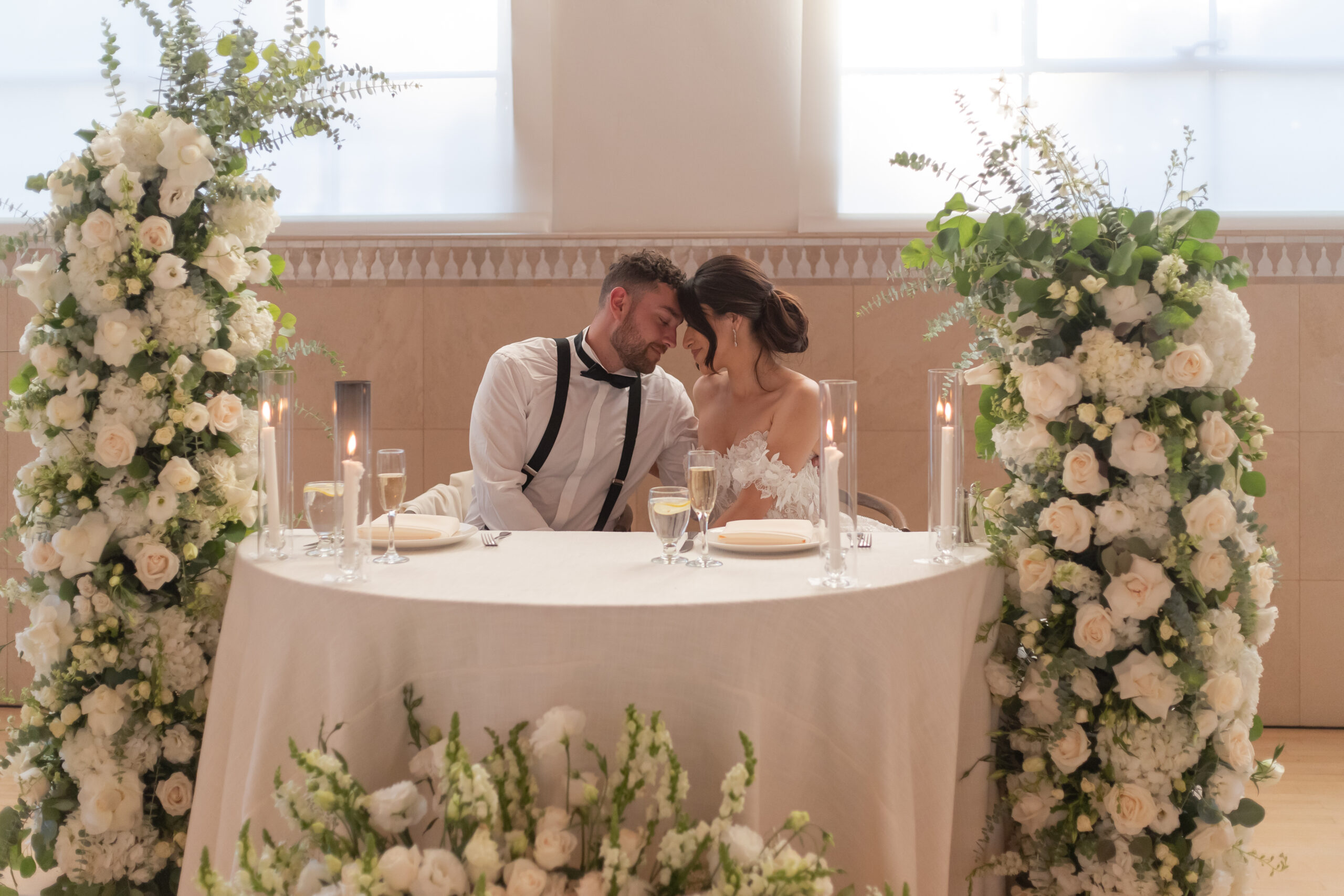 Bride and groom sitting at a neutral white sweetheart table adorned with freestanding white and greenery floral arrangements.