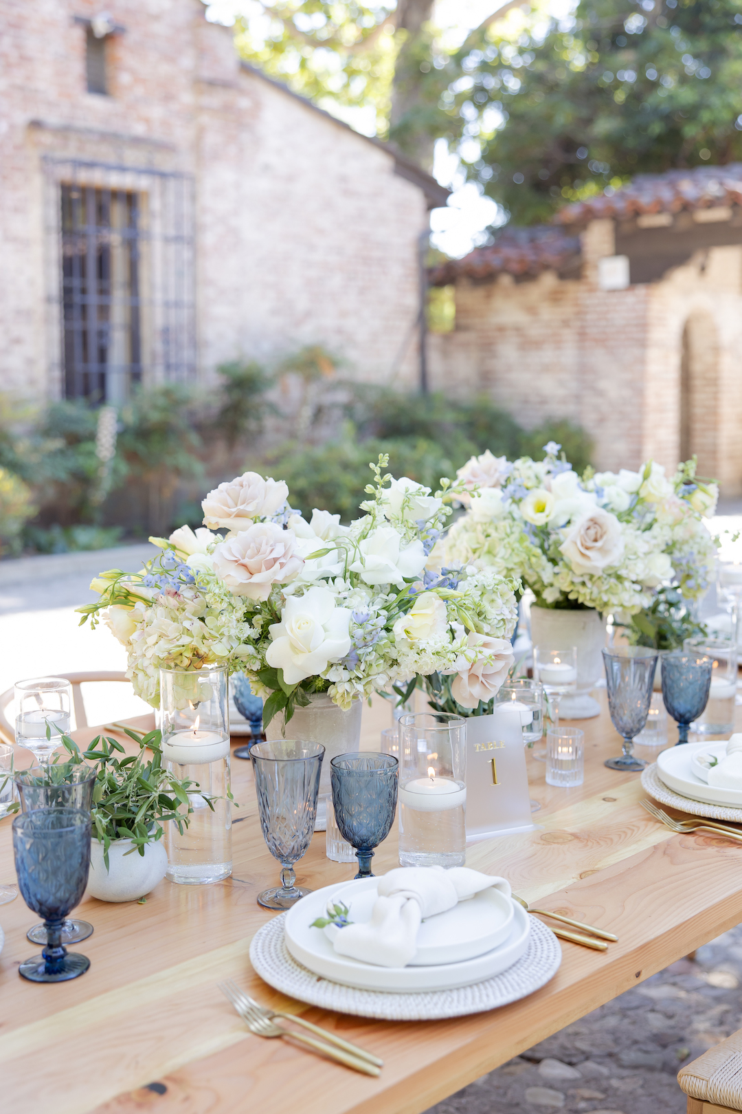Elegant wedding table decor with glass candles, blue glassware, floral arrangements, ceramic plates, and gold-finished silverware. 