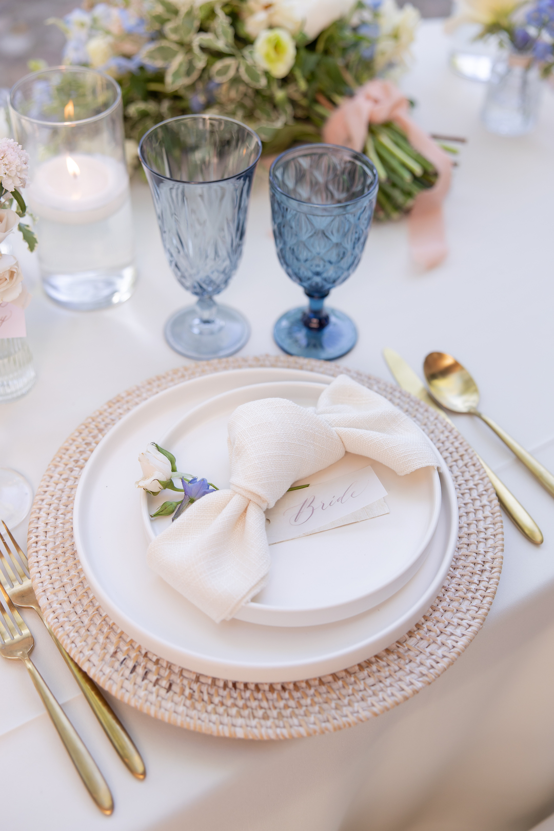 Wedding place setting with ceramic plates, wicker chargers, bow-tied linen napkins and gold flatware. 