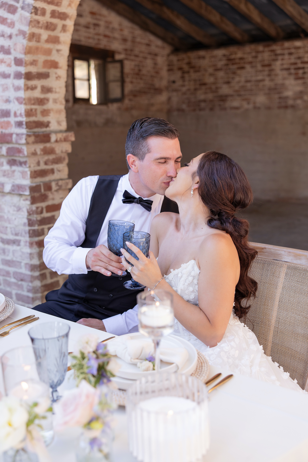 Bride and groom clinking blue stained glass cups and sharing a kiss at their wedding sweetheart table. 