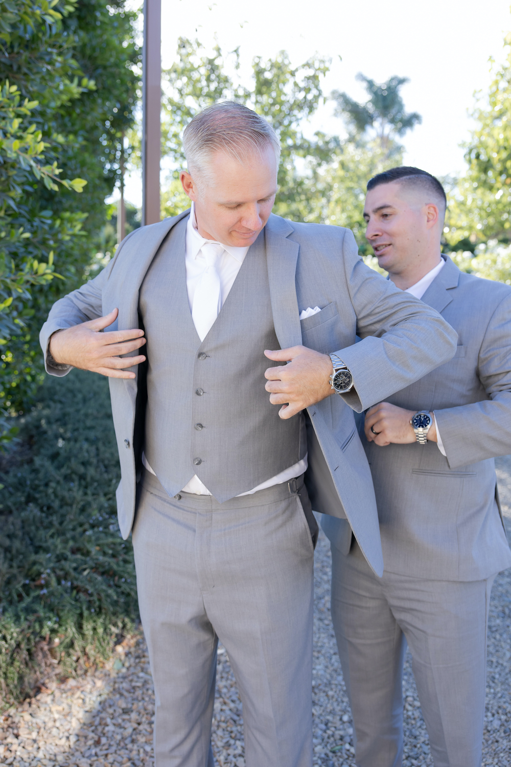 Groom in a classic grey suit putting on his jacket with the assistance of his groomsman.